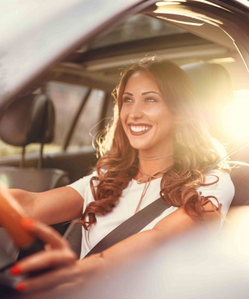 Beautiful young happy smiling woman driving her new car at sunset.